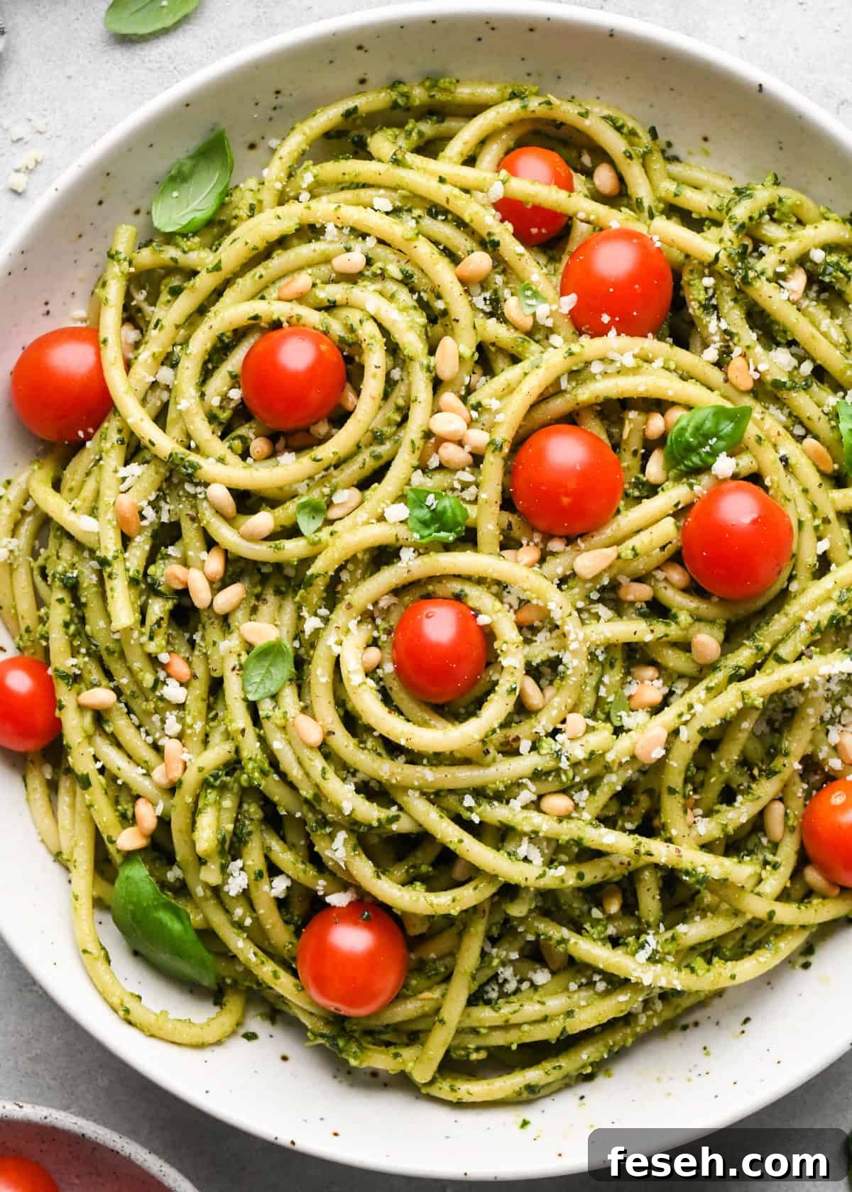 pesto pasta in a ceramic bowl with cherry tomatoes and a silver fork, garnished with toasted pine nuts, parmesan cheese and fresh basil on a gray surface