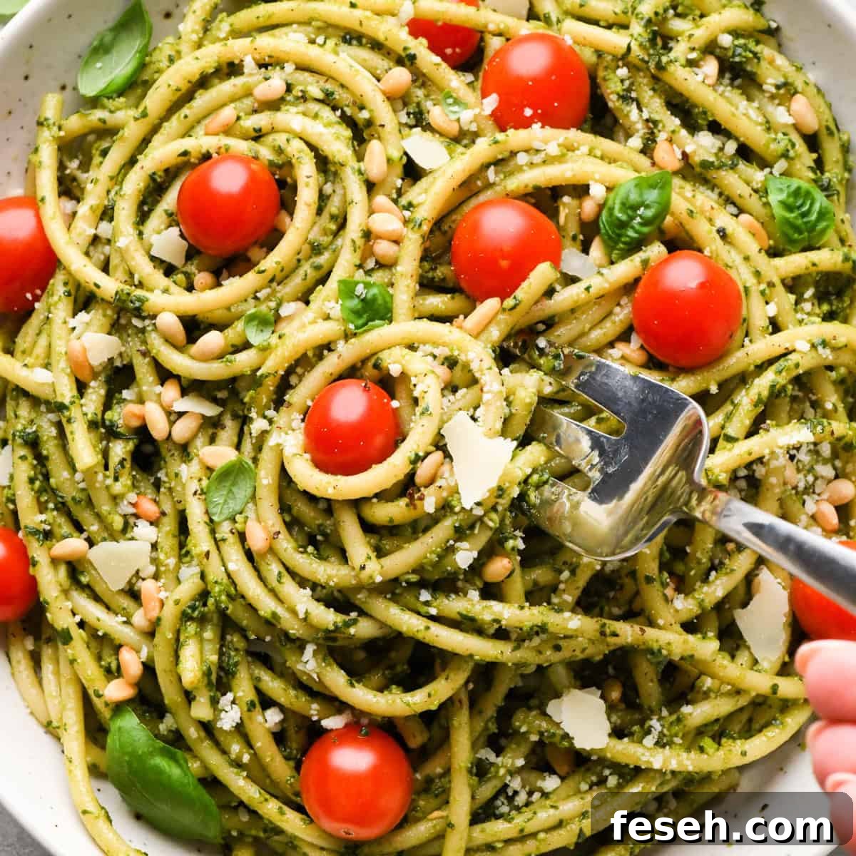 a fork taking a bite of pesto pasta in a ceramic bowl with cherry tomatoes and a silver fork, garnished with toasted pine nuts, parmesan cheese and fresh basil on a gray surface surrounded by basil, pine nuts, tomatoes and a striped towel.
