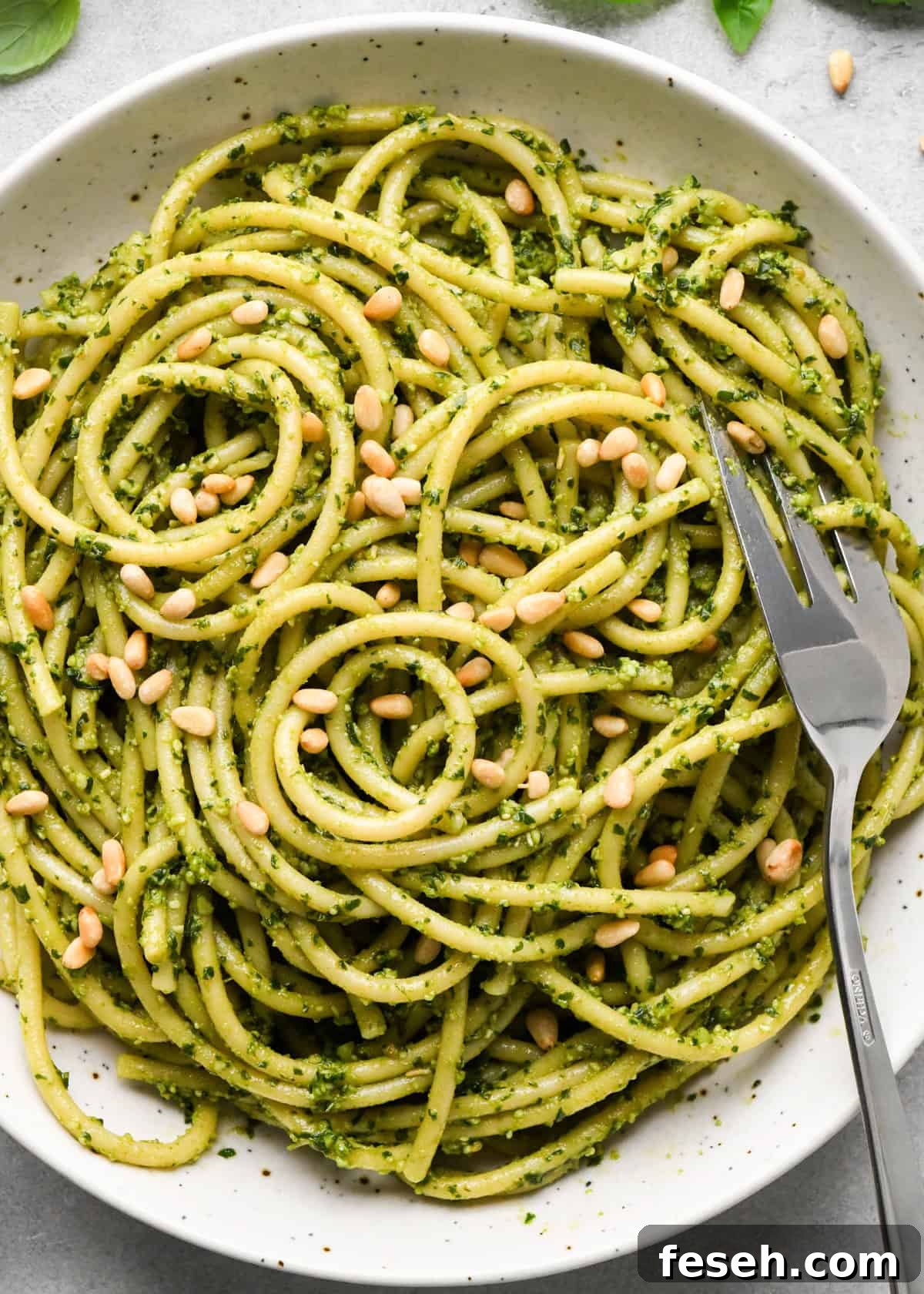 close up photo of pesto pasta in a ceramic bowl with a silver fork, garnished with toasted pine nuts on a gray surface