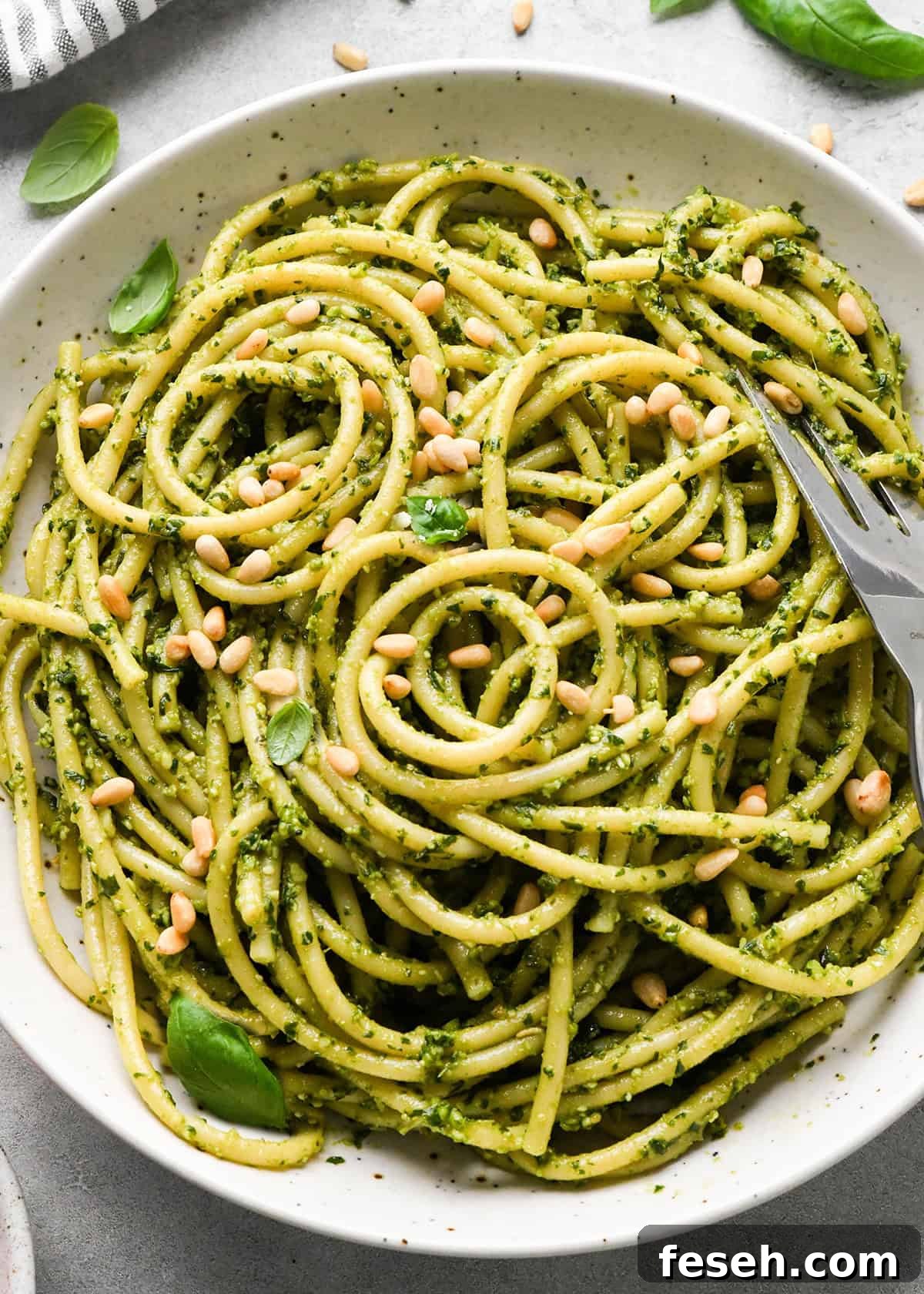 close up photo of pesto pasta in a ceramic bowl with a silver fork, garnished with basil and toasted pine nuts on a gray surface