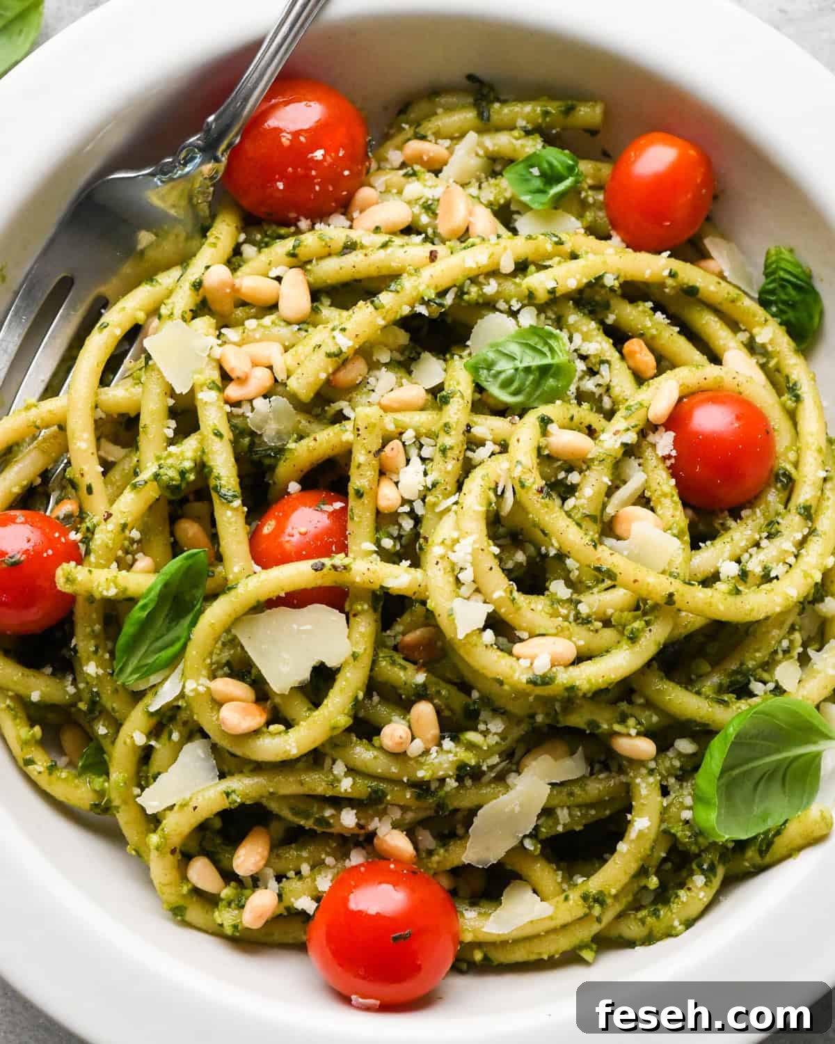 pesto pasta in a white bowl with cherry tomatoes and a silver fork, garnished with toasted pine nuts, parmesan cheese and fresh basil