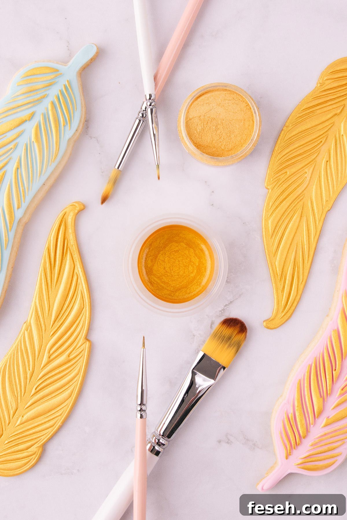 A small plastic container of edible gold paint, surrounded by pink and white handled paintbrushes, and feather-shaped cookies painted with gold paint. 