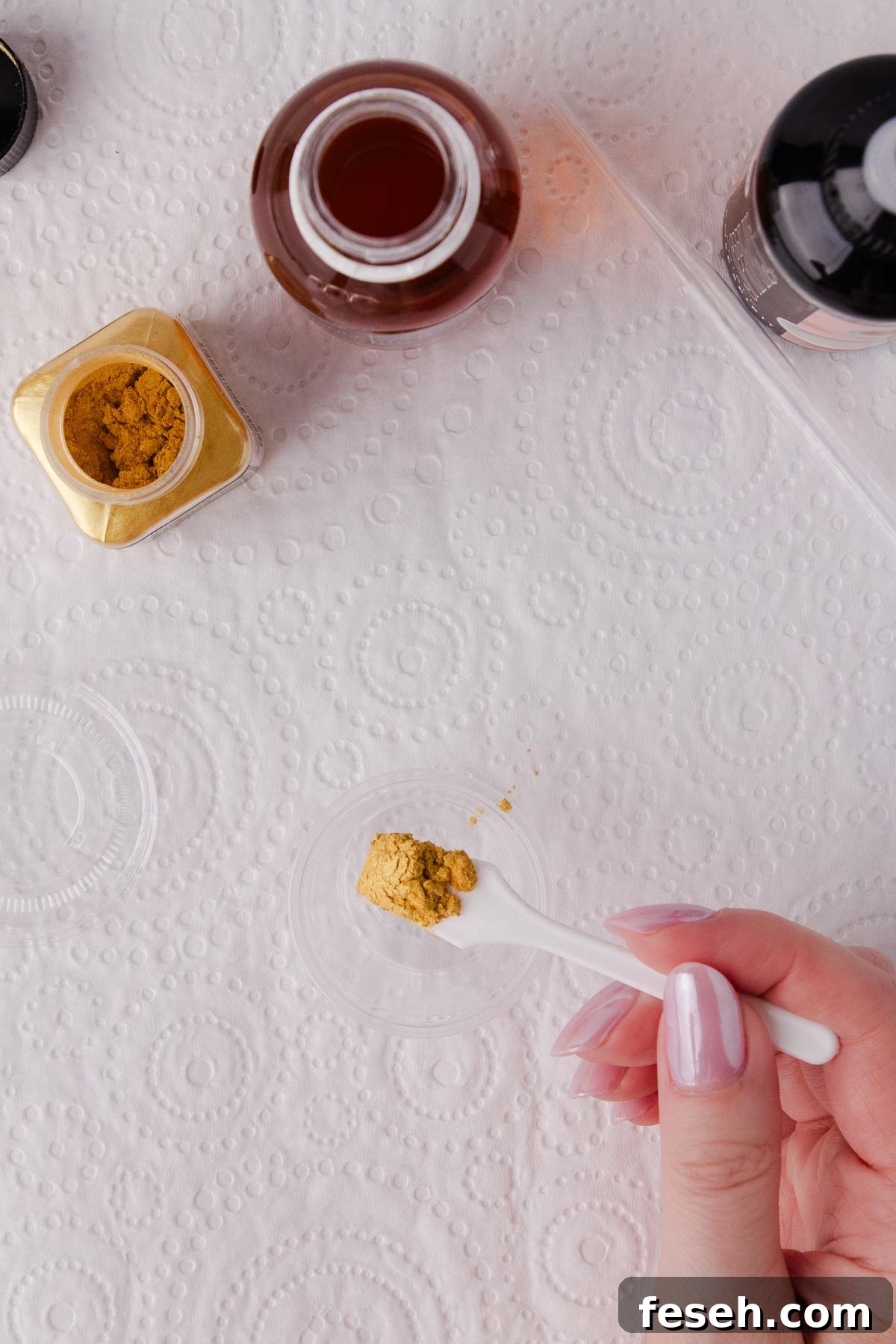 A small white spoon with about 1/4 teaspoon of gold edible lustre powder over a small plastic container.