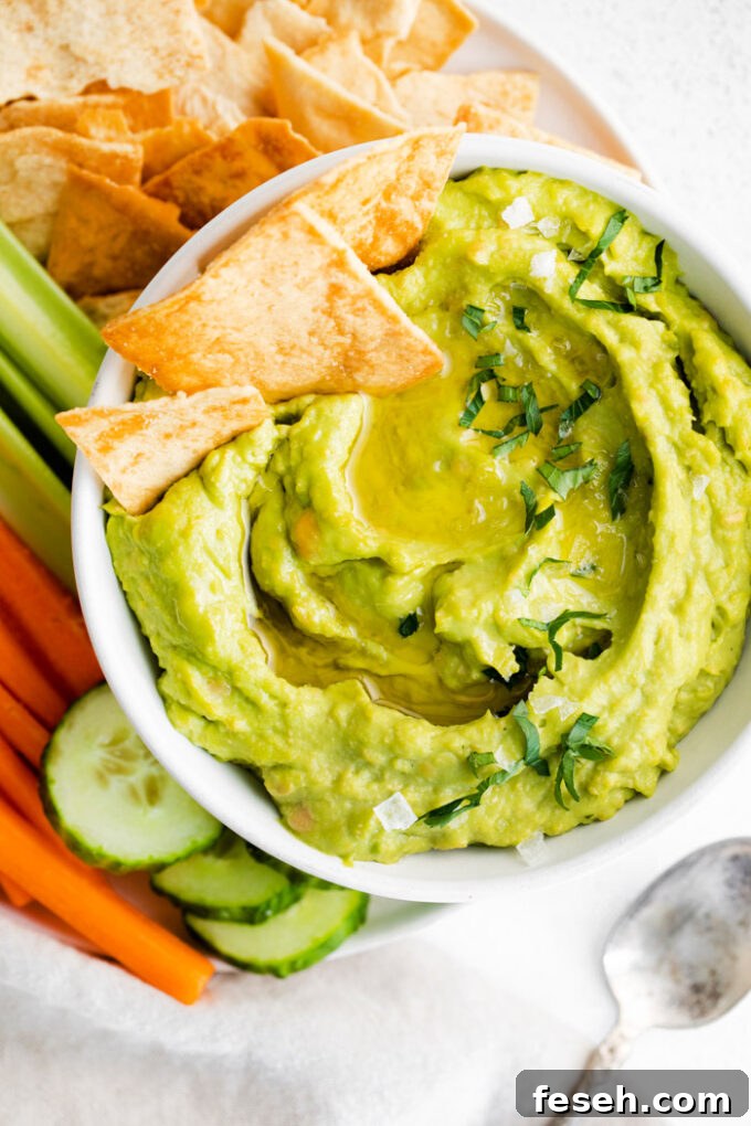 Creamy Avocado Hummus Recipe 2 white bowl filled with green avocado hummus and surrounded by chips and veggies. a few chips sticking out of the hummus
