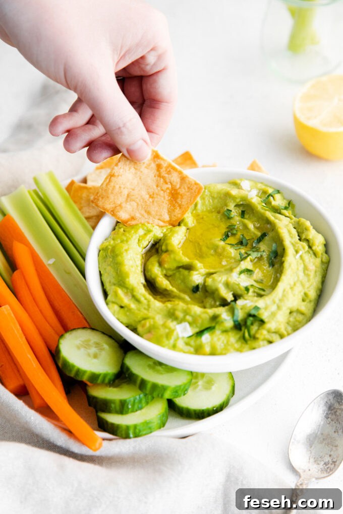 Creamy Avocado Hummus Recipe 5 white bowl filled with green hummus with a hand holding a chip being dipped in the hummus. fresh veggies around the bowl