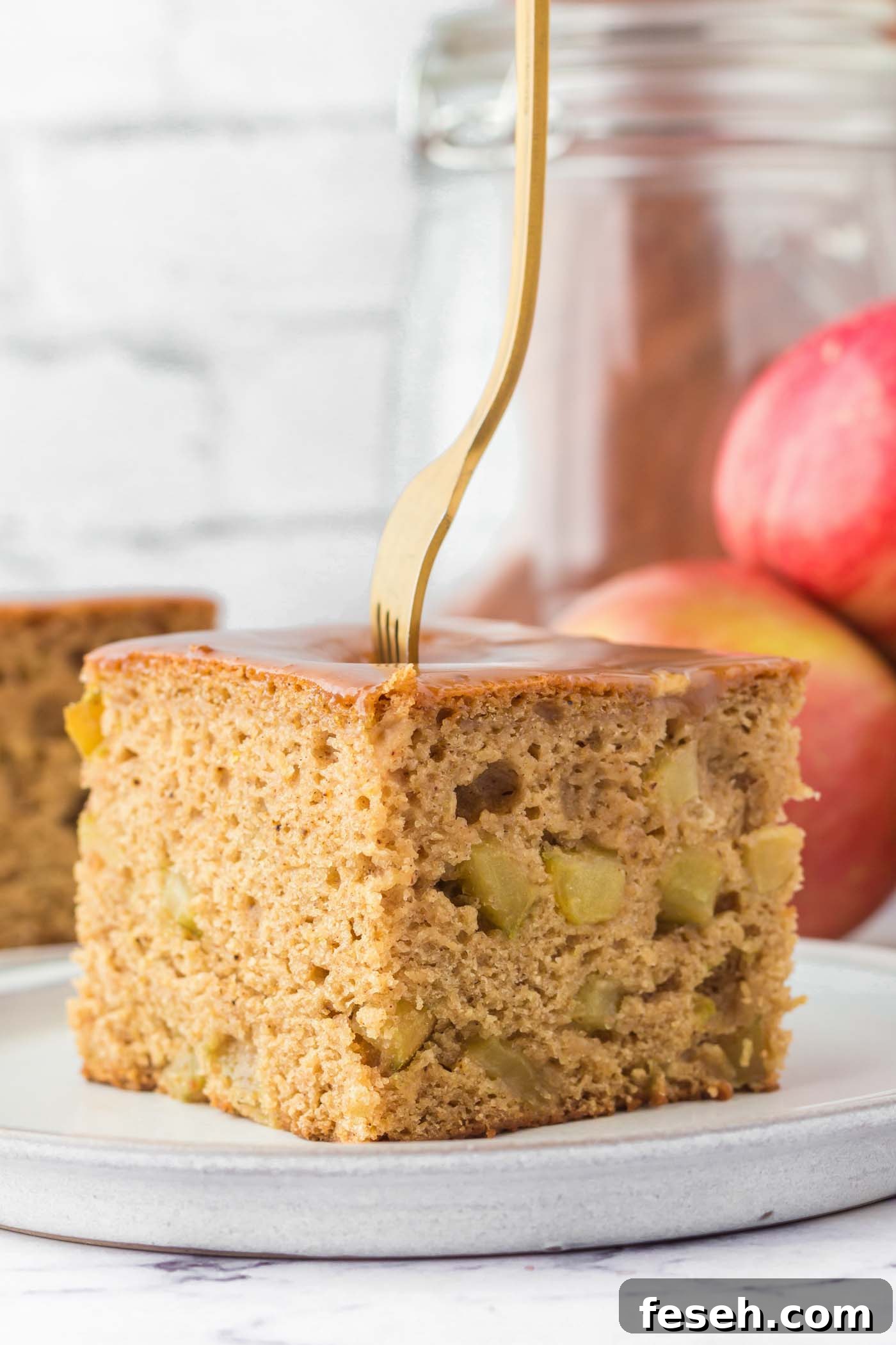 Moist Vegan Apple Cake with Cinnamon Glaze 2 A large square of vegan apple cake on a plate. The cake is topped with a caramel glaze, there's a fork stuck in the cake and you can see some pieces of diced apple in the cake.