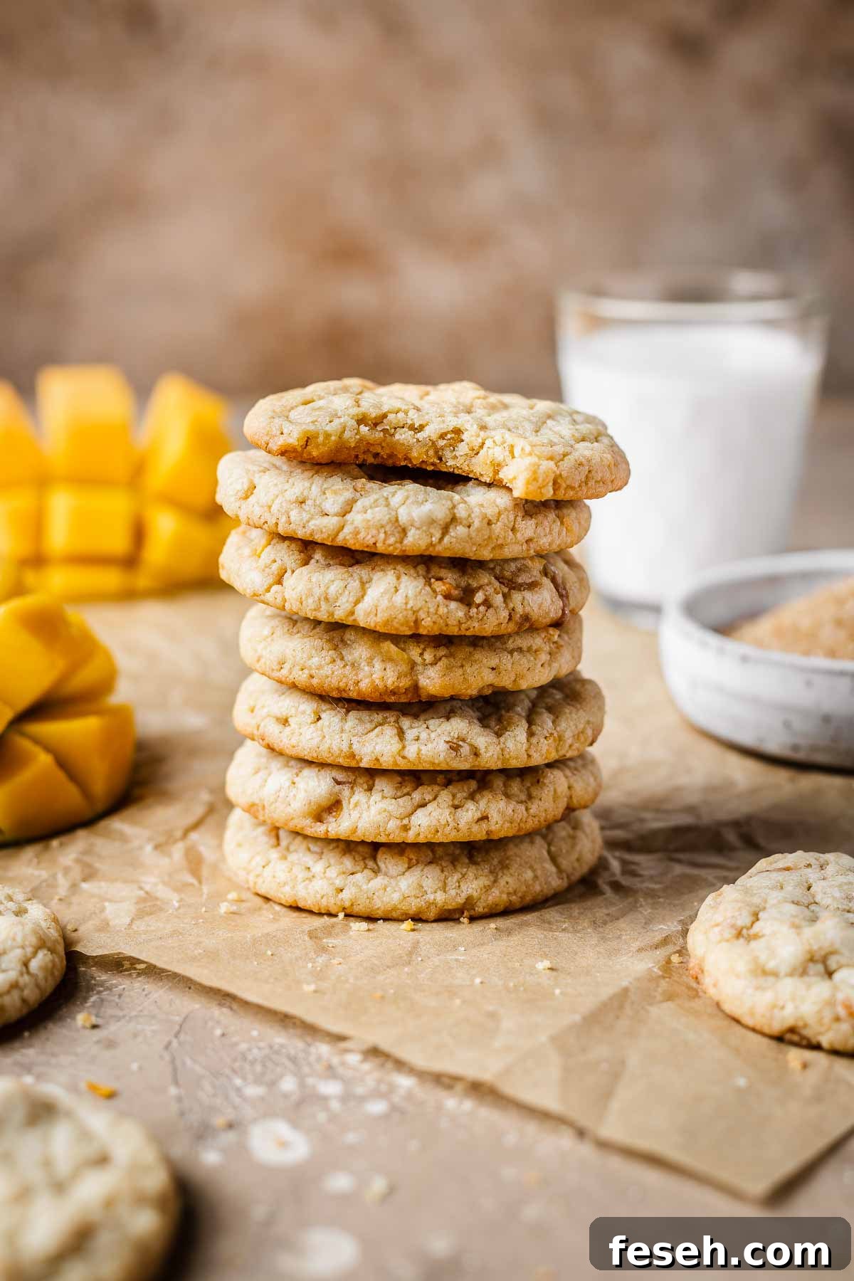 Tropical Mango Cookies with Coconut Glaze 14 A stack of seven mango cookies on brown parchment paper with a glass of milk and sliced mangos in the background.
